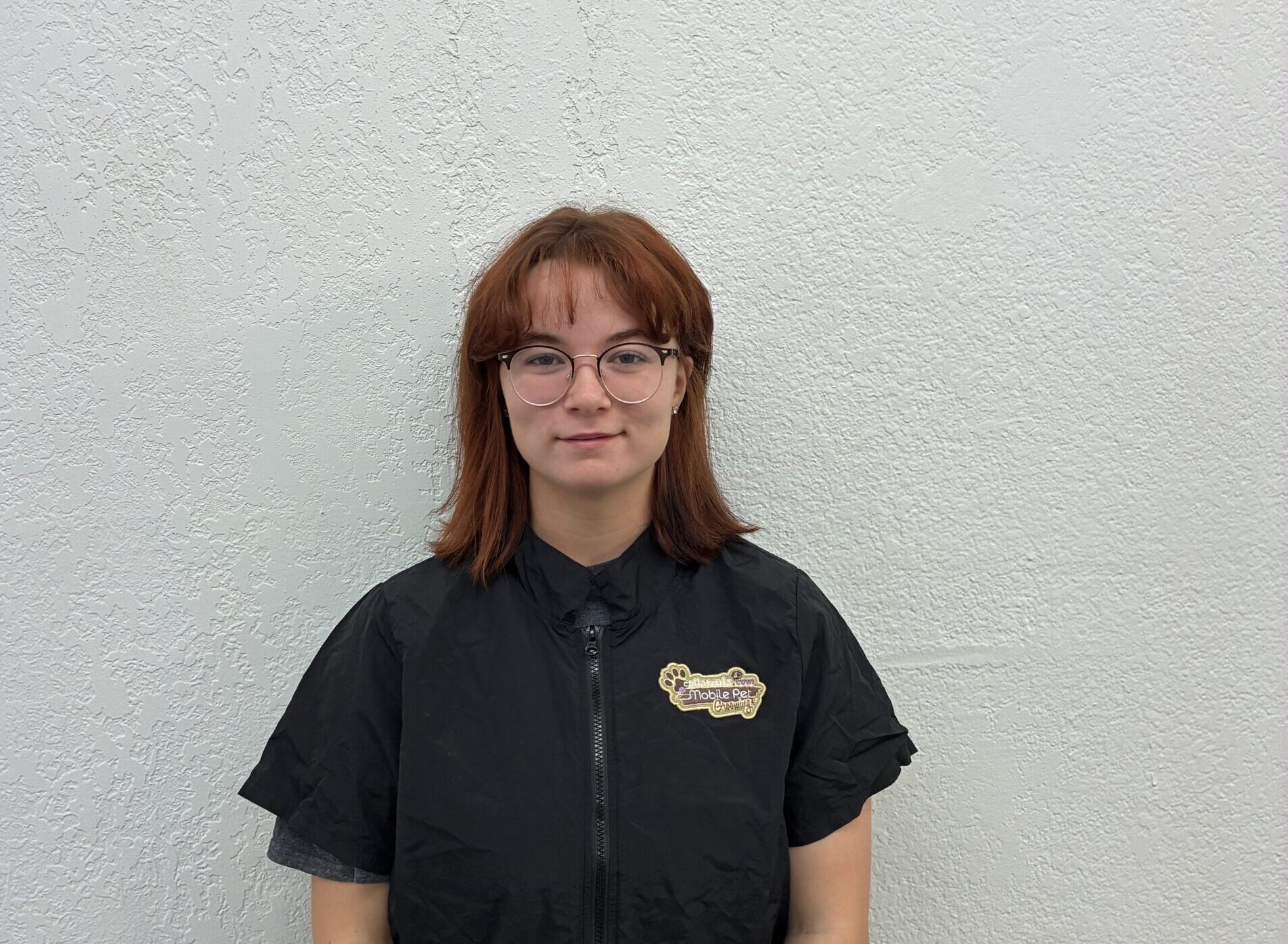 Young woman with red hair and round glasses stands against a pale, textured wall, wearing a black zip-up jacket with a badge on the left chest area.